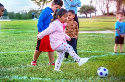 children gathering around a young girl kicking a soccer ball in a soccer field