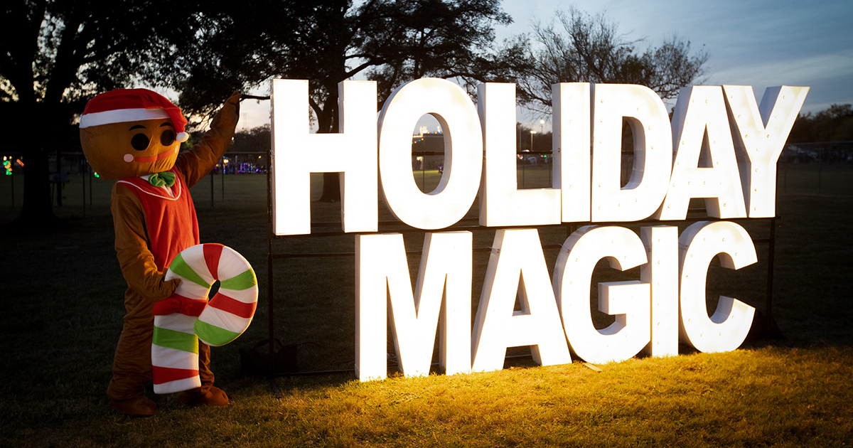 A gingerbread man mascot stands next to a large neon Holiday Magic sign.
